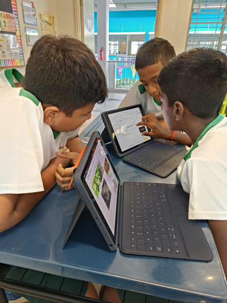 Three boys in white and green uniforms huddle around two tablets with keyboards at a classroom desk.